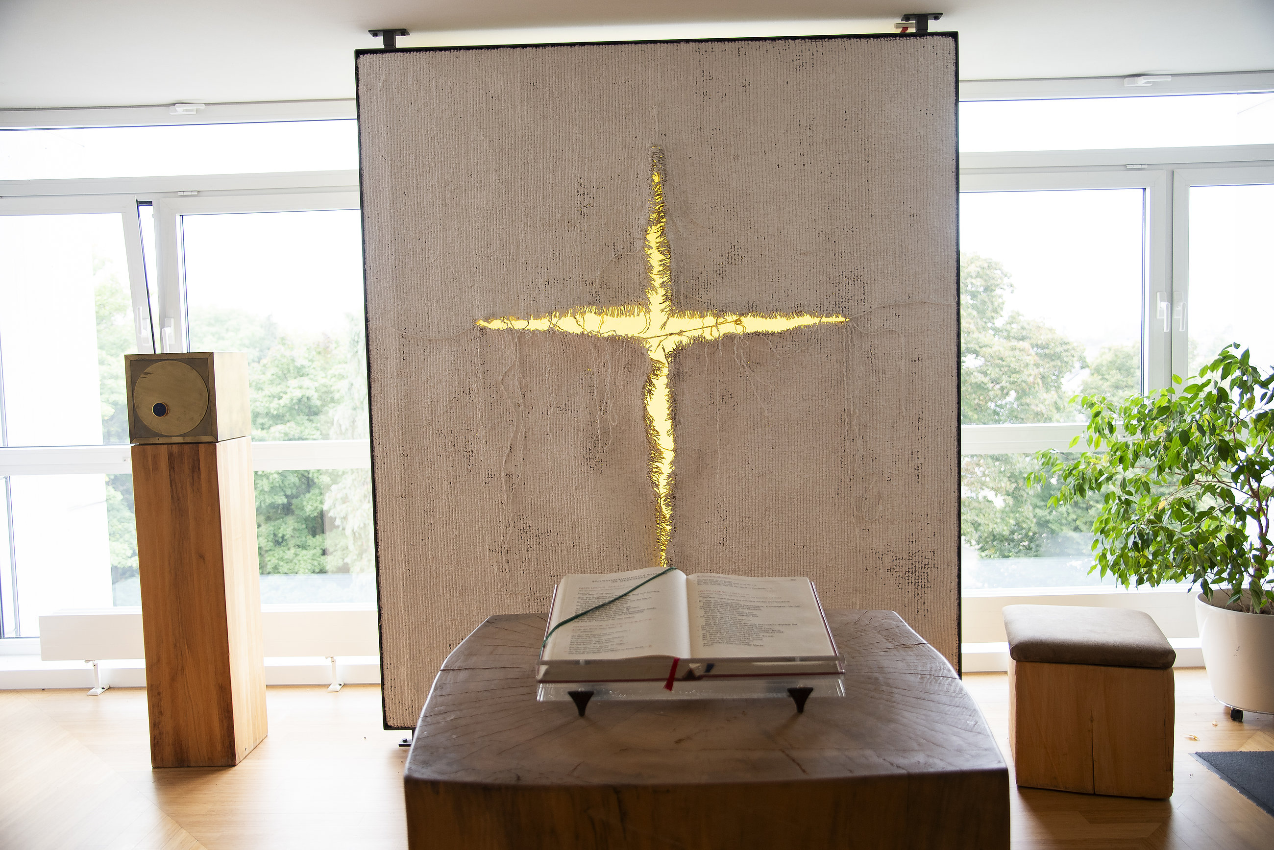 Offener Bibelband auf hölzernem Lesepult vor Wand mit goldenem Kreuz in hellem Raum mit Fenstern und Pflanze
