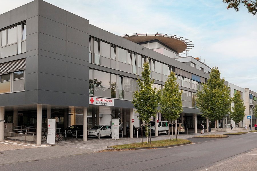 Moderne, mehrstöckige Gebäude mit großen Fenstern und einem begrünten Dach. Vor dem Gebäude stehen mehrere Bäume und Autos. Eine Straße verläuft entlang der Vorderseite.
