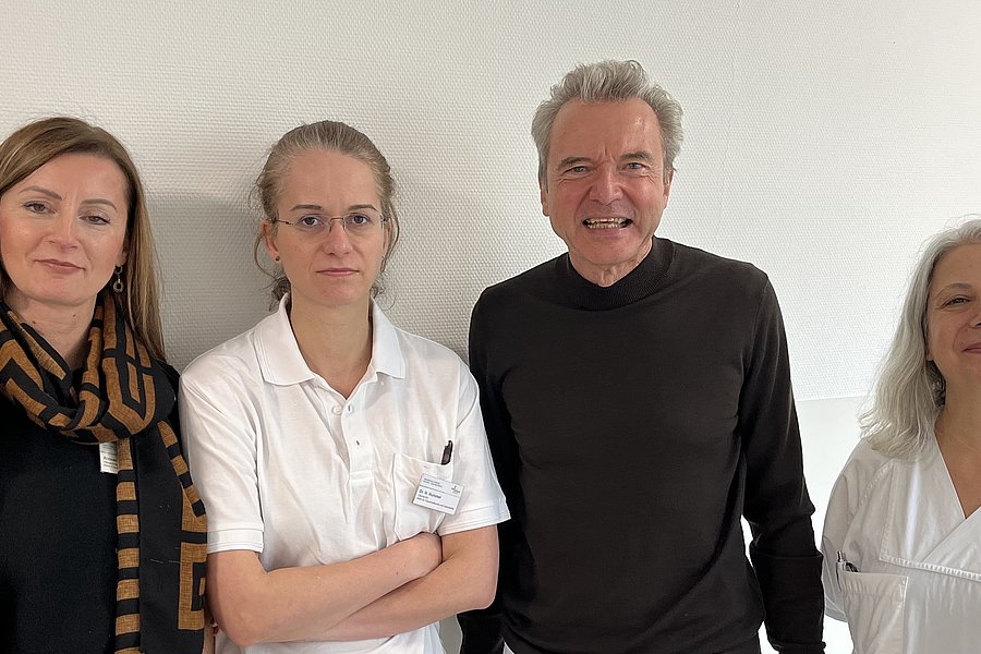 Vier Personen stehen nebeneinander vor einer hellen Wand, zwei Frauen links tragen helle Oberteile, ein Mann in der Mitte trägt ein dunkles Oberteil, eine Frau rechts trägt ein weißes Oberteil