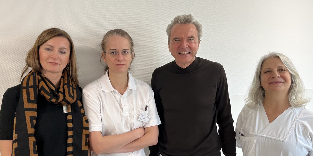 Vier Personen stehen nebeneinander vor einer hellen Wand, zwei Frauen links tragen helle Oberteile, ein Mann in der Mitte trägt ein dunkles Oberteil, eine Frau rechts trägt ein weißes Oberteil