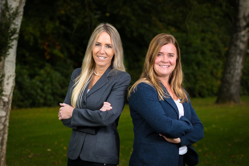Zwei Frauen stehen nebeneinander im Freien, beide mit verschränkten Armen. Die Frau links trägt einen grauen Blazer, die Frau rechts einen blauen Blazer.