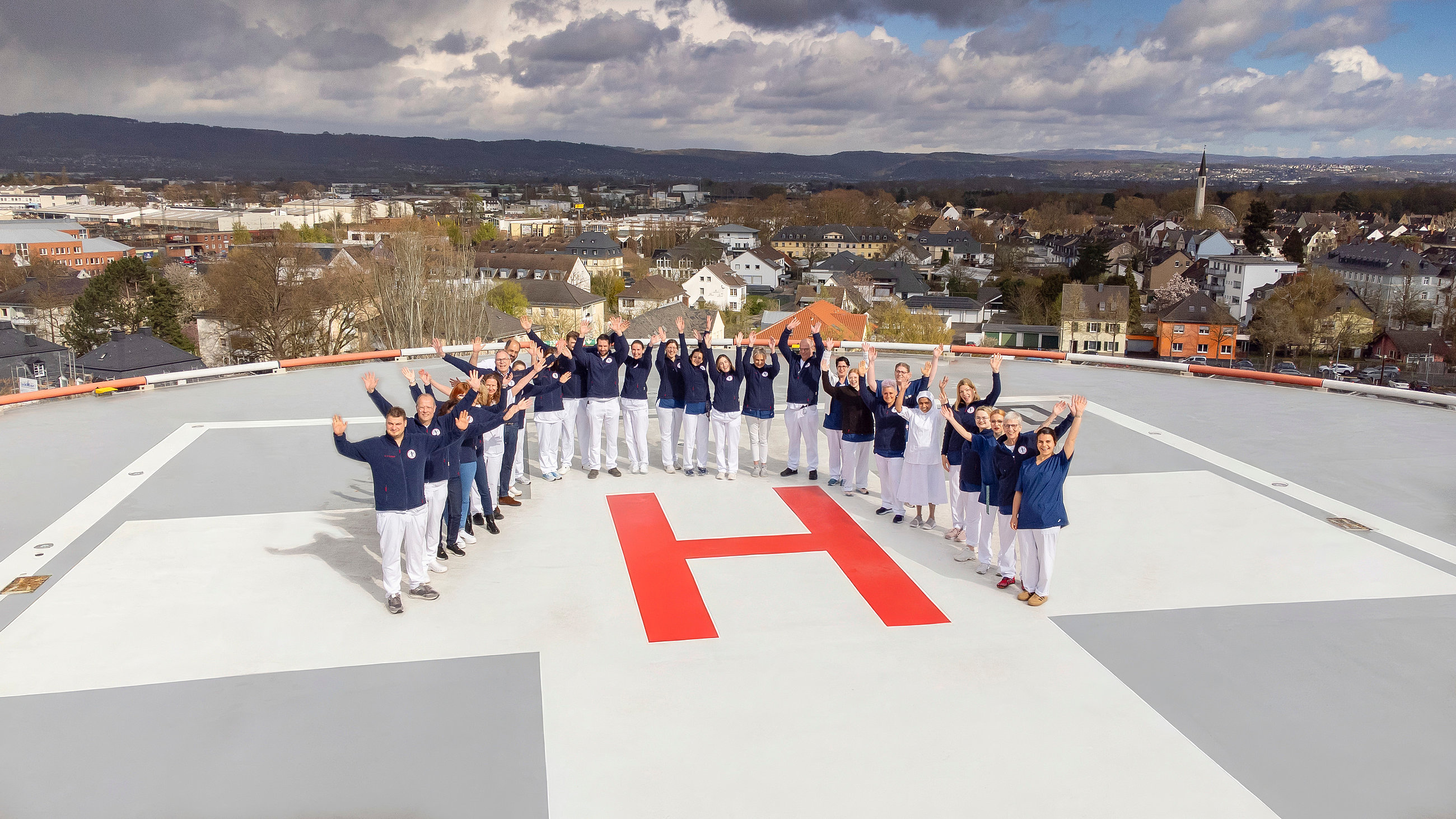 Gruppe von Menschen in blauen Oberteilen und weißen Hosen steht auf einem Hubschrauberlandeplatz in Form eines H und hebt die Arme in die Luft