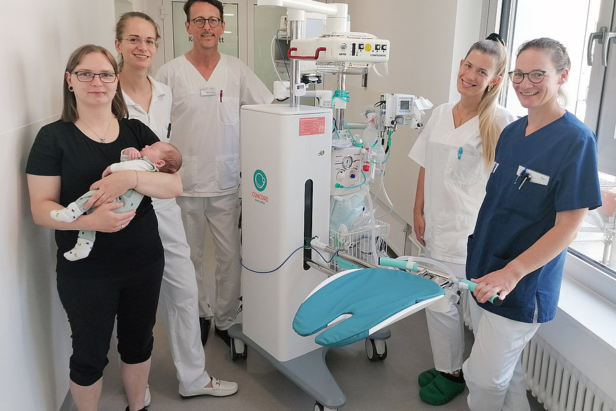 Sechs Personen in medizinischer Kleidung stehen in einem Raum. Eine Person hält ein Baby, während eine andere ein medizinisches Gerät mit einer blauen Liege präsentiert.