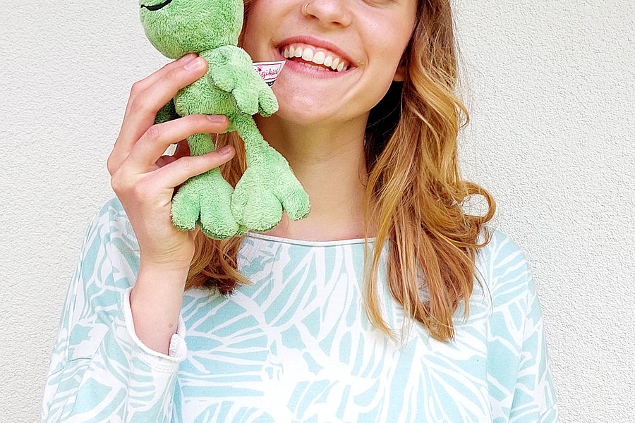 Eine Person mit langen, lockigen Haaren hält ein grünes Stofftier in Form eines Frosches in der Hand. Sie trägt ein hellblaues Oberteil mit einem Blattmuster.