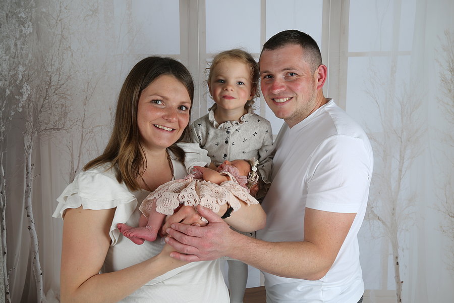 Eine Familie steht zusammen, die Mutter hält ein Baby in einem rosa Kleid, der Vater steht daneben. Ein Kind mit blonden Haaren steht zwischen ihnen.