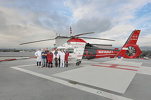 Gruppe von medizinischem Personal in weißer und roter Kleidung steht neben einem Rettungshubschrauber mit der Aufschrift "NOTARZT" auf einem Hubschrauberlandeplatz auf einem Dach