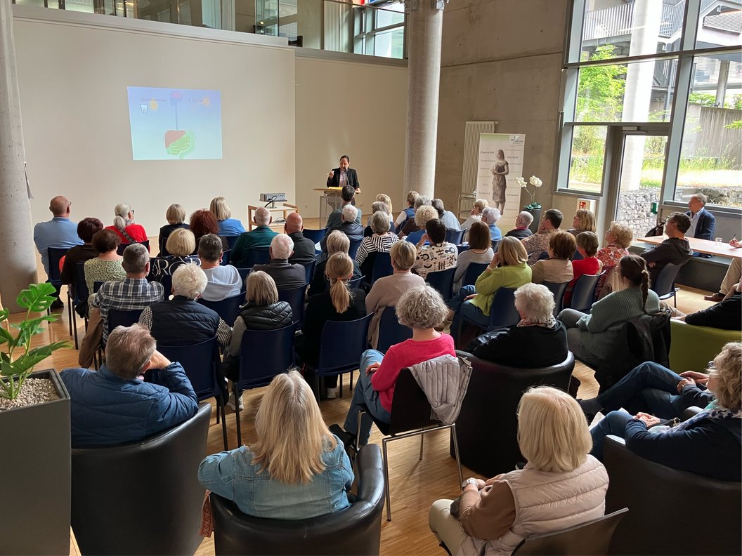 Publikum sitzt in einem modernen Raum und hört einem Vortragenden zu, der vor einer Leinwand steht