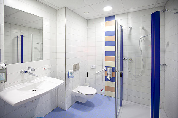 Barrierefreies Badezimmer mit Wand-WC, Waschbecken, großer Spiegel und ebenerdiger Dusche mit blauen Akzenten