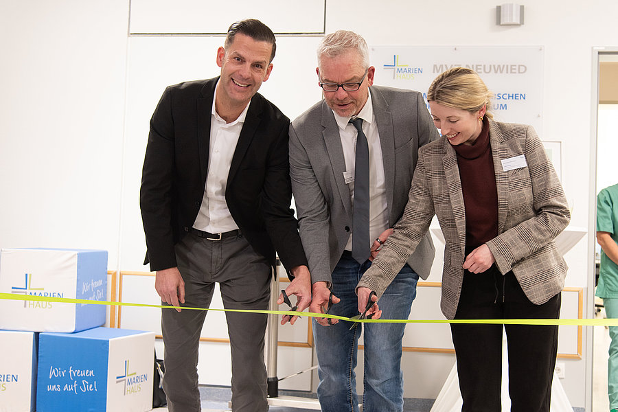 Drei Personen in Businesskleidung schneiden gemeinsam ein gelbes Band durch, im Hintergrund stehen Kartons und eine Person in grüner Arbeitskleidung.