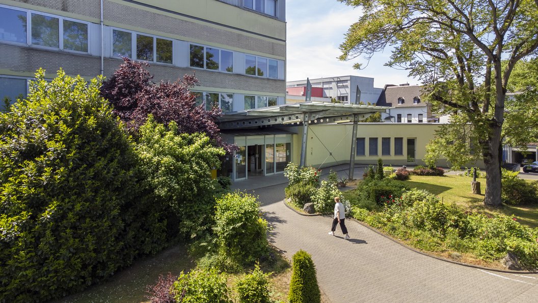 Gebäudekomplex mit mehreren Stockwerken, umgeben von Bäumen und Sträuchern, auf einem gepflasterten Weg geht eine Person mit Tasche