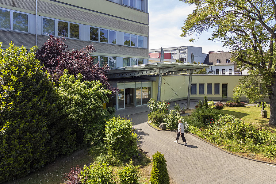 Gebäudekomplex mit mehreren Stockwerken, umgeben von Bäumen und Sträuchern, auf einem gepflasterten Weg geht eine Person mit Tasche