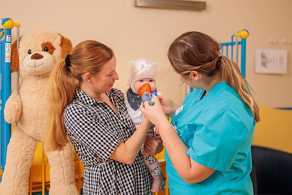 Kinderärztin in blauem Kittel untersucht Baby, das von Frau in kariertem Hemd gehalten wird, großer Plüschhund im Hintergrund