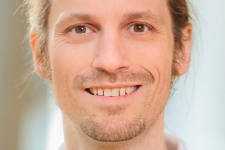 Frank Martin Kügler trägt ein weißes Polohemd und hat lange, blonde Haare, die zu einem Zopf gebunden sind. Der Hintergrund ist unscharf.