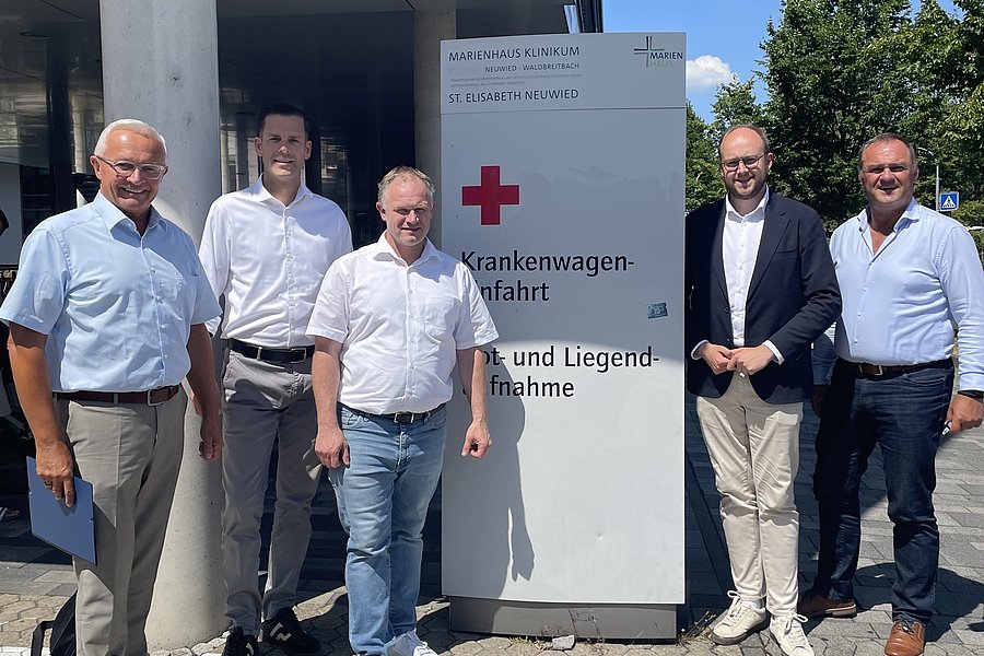 Fünf Männer in Hemden und Hosen stehen vor einem Schild mit rotem Kreuz und Text vor einem Gebäude unter blauem Himmel