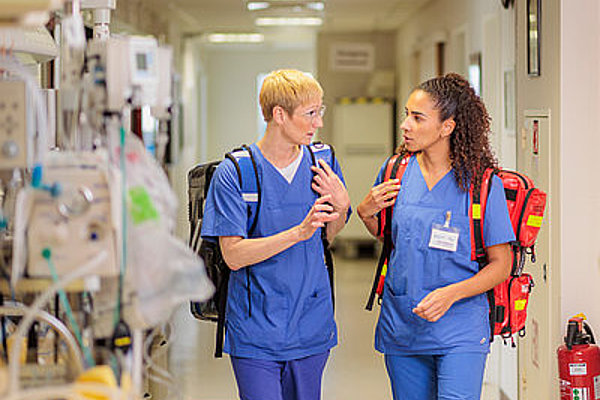 Zwei Pflegekräfte in blauen Kitteln mit Rucksäcken sprechen in einem Krankenhausflur