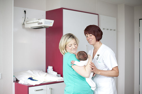 Zwei Frauen in medizinischer Umgebung, eine hält ein Baby, die andere berührt sanft den Arm der Frau mit dem Baby