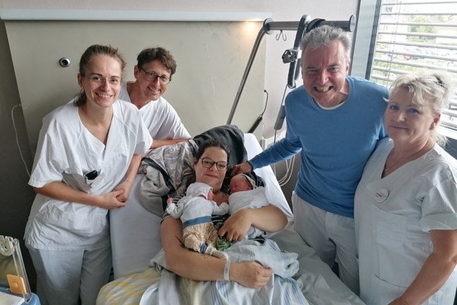 Eine Frau hält ein Neugeborenes in einem Krankenhausbett, umgeben von fünf Personen in weißen Kitteln. Die Personen stehen und sitzen nahe bei ihr.