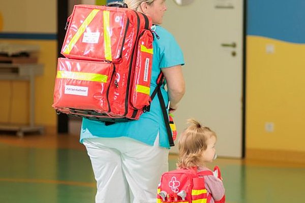 Erwachsene mit rotem Rettungsrucksack und Kind mit kleinem rotem Rucksack in einem Flur mit gelben und blauen Wänden
