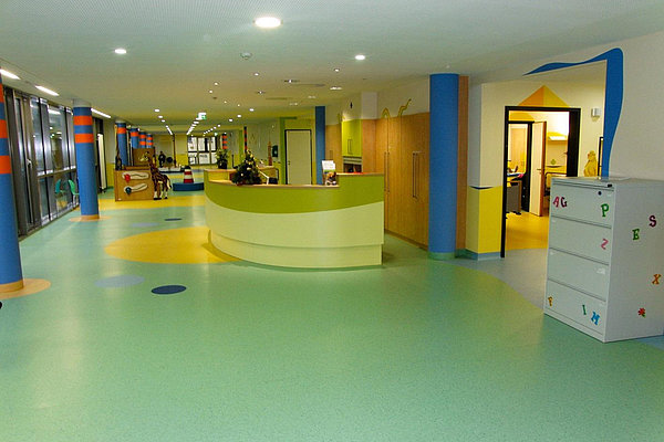 Der Flur der Klinik für Kinder und Jugendmedizin ist hell und farbenfroh gestaltet. Ein geschwungener Empfangstresen steht in der Mitte, links sorgen große Fenster für viel Licht.
