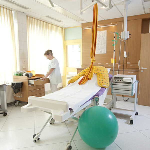 Krankenhauszimmer mit Behandlungsliege, grünem Gymnastikball, medizinischem Gerät und einer Person in weißer Kleidung am Schreibtisch