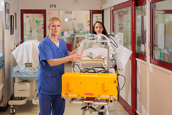 Zwei medizinische Fachkräfte schieben einen Inkubator mit einem Neugeborenen durch einen Krankenhausflur