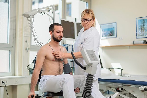 Medizinisches Personal führt Belastungs-EKG bei einem Patienten in der Kardiologie durch, Patient sitzt auf Ergometer mit Elektroden am Oberkörper