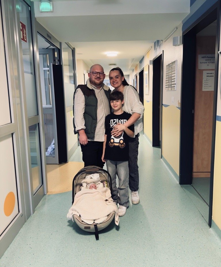 Drei Personen stehen in einem Flur, ein Kind hält einen Kinderwagen mit einer Decke darin