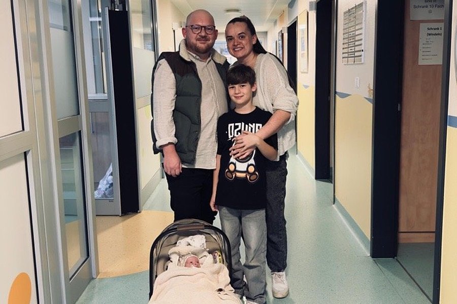 Drei Personen stehen in einem Flur, ein Kind hält einen Kinderwagen mit einer Decke darin