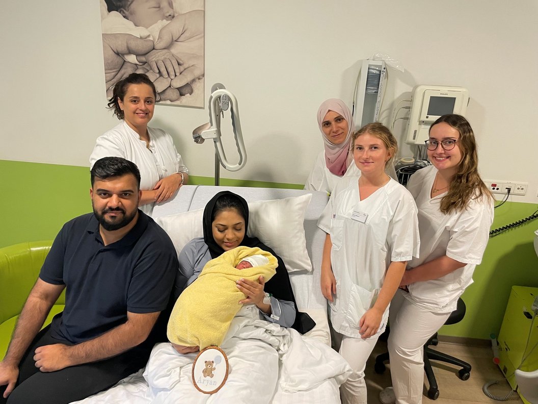 Gruppe von sechs Personen in einem Krankenhauszimmer, eine Frau hält ein in eine gelbe Decke gewickeltes Baby auf dem Schoß, umgeben von fünf weiteren Personen, von denen vier weiße Pflegekleidung tragen
