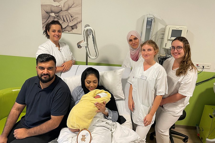 Gruppe von sechs Personen in einem Krankenhauszimmer, eine Frau hält ein in eine gelbe Decke gewickeltes Baby auf dem Schoß, umgeben von fünf weiteren Personen, von denen vier weiße Pflegekleidung tragen