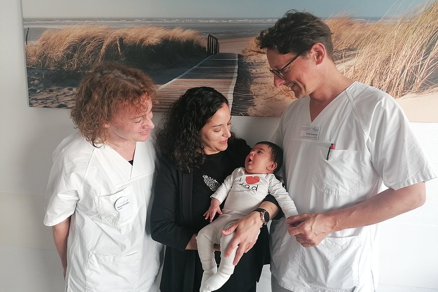 Zwei Personen in medizinischer Kleidung stehen zusammen. Eine dritte Person in der Mitte hält ein Baby in einem weißen Outfit. Im Hintergrund ist ein Bild von einer Landschaft mit Strand und Dünen zu sehen.