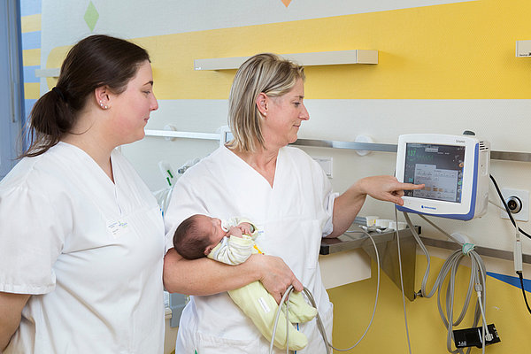 Zwei Pflegekräfte in weißen Kitteln bedienen ein medizinisches Überwachungsgerät, eine hält ein schlafendes Baby in gelber Decke