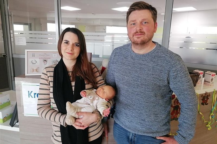 Junges Paar steht am Empfang des Kreißsaals, die Frau hält ein schlafendes Baby im Arm, beide blicken in die Kamera