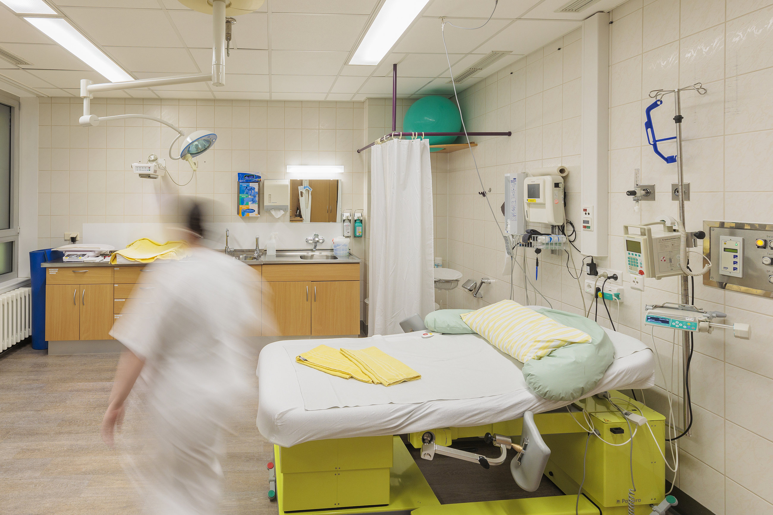 Krankenhauszimmer mit Bett, medizinischen Geräten und einer verschwommenen Person in weißer Kleidung