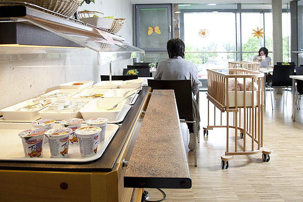 Buffet mit Joghurtbechern und Schalen in einem hellen Raum, zwei Personen sitzen an Tischen, ein Kinderbett auf Rollen steht daneben