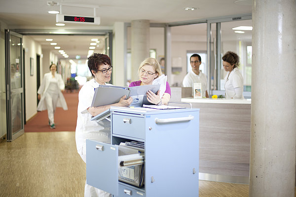 Zwei medizinische Fachkräfte besprechen Unterlagen an einem blauen Rollwagen in einem Krankenhausflur, im Hintergrund weitere Personen am Empfang