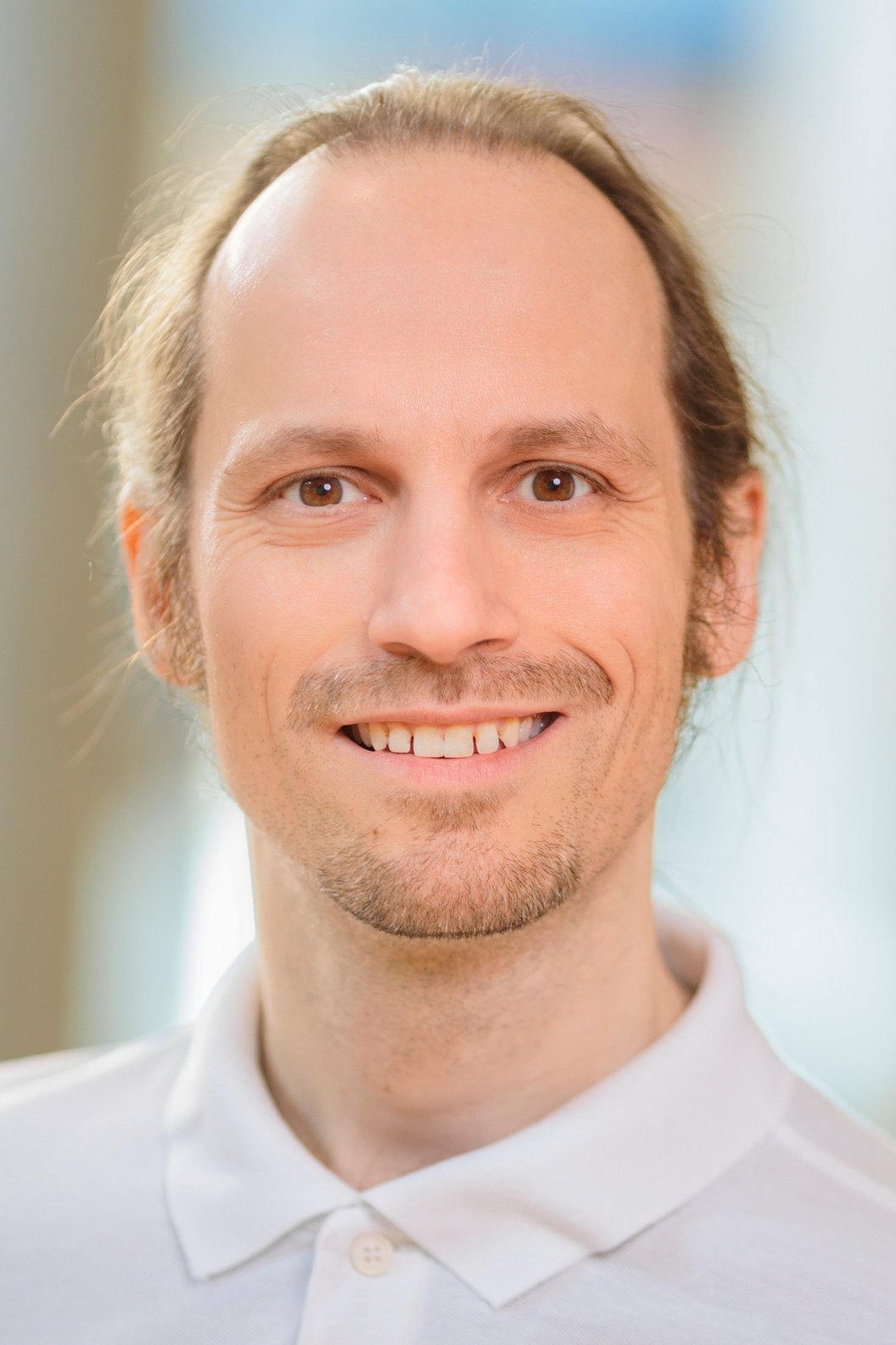 Frank Martin Kügler trägt ein weißes Polohemd und hat lange, blonde Haare, die zu einem Zopf gebunden sind. Der Hintergrund ist unscharf.