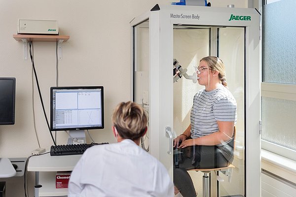 Frau führt Lungenfunktionstest in Jaeger MasterScreen Body Kabine durch, während medizinisches Personal am Computer die Messwerte überwacht