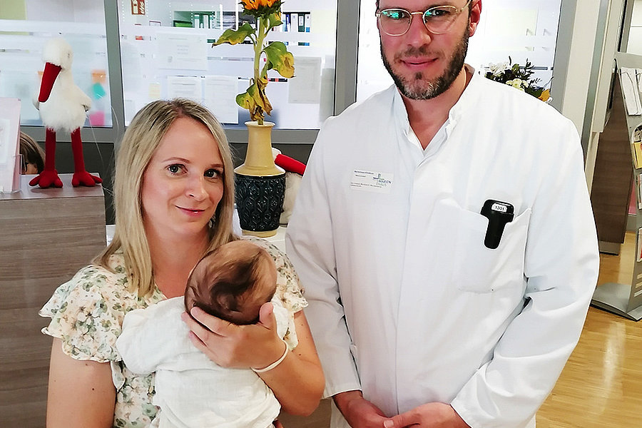 Eine Frau hält ein Baby in ihren Armen, während ein Mann in einem weißen Kittel neben ihr steht. Im Hintergrund sind Pflanzen und Dekorationen sichtbar.