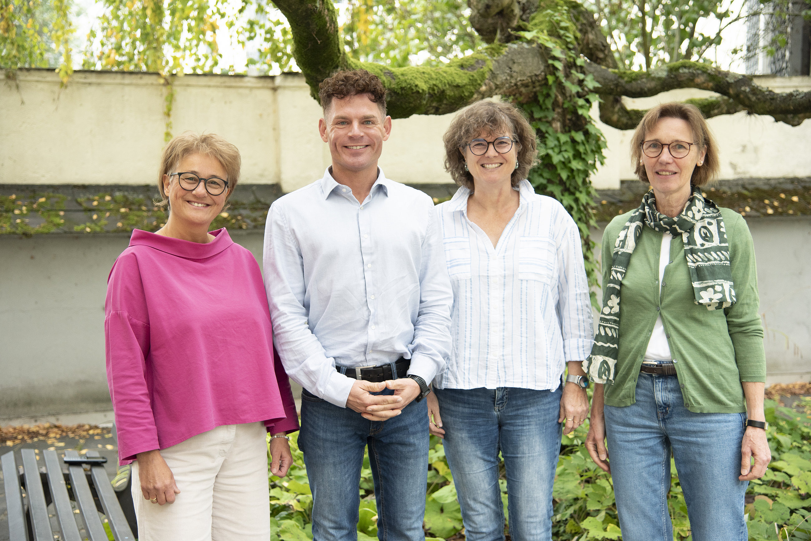 Vier Personen stehen nebeneinander vor einem Baum und einer Mauer, zwei Frauen tragen Pullover, ein Mann ein Hemd, eine Frau eine Jacke mit Schal