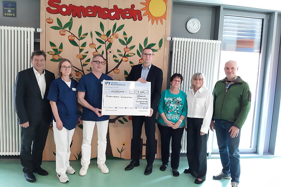 Gruppe von sieben Personen steht vor einer Wand mit einem Baum und der Aufschrift 'Sonnenschein', zwei Personen halten einen großen symbolischen Scheck