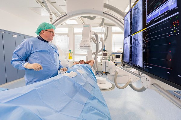 Dr. Hügl führt Herzkatheter-Untersuchung bei Patientin im OP-Saal mit Monitoren und medizinischer Ausrüstung durch