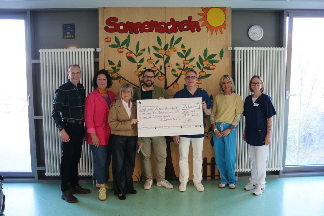 Gruppe von sieben Personen steht in einem Raum vor einer Wand mit einem großen bunten Wandbild mit der Aufschrift 'Sonnenschein' und hält einen großen symbolischen Scheck