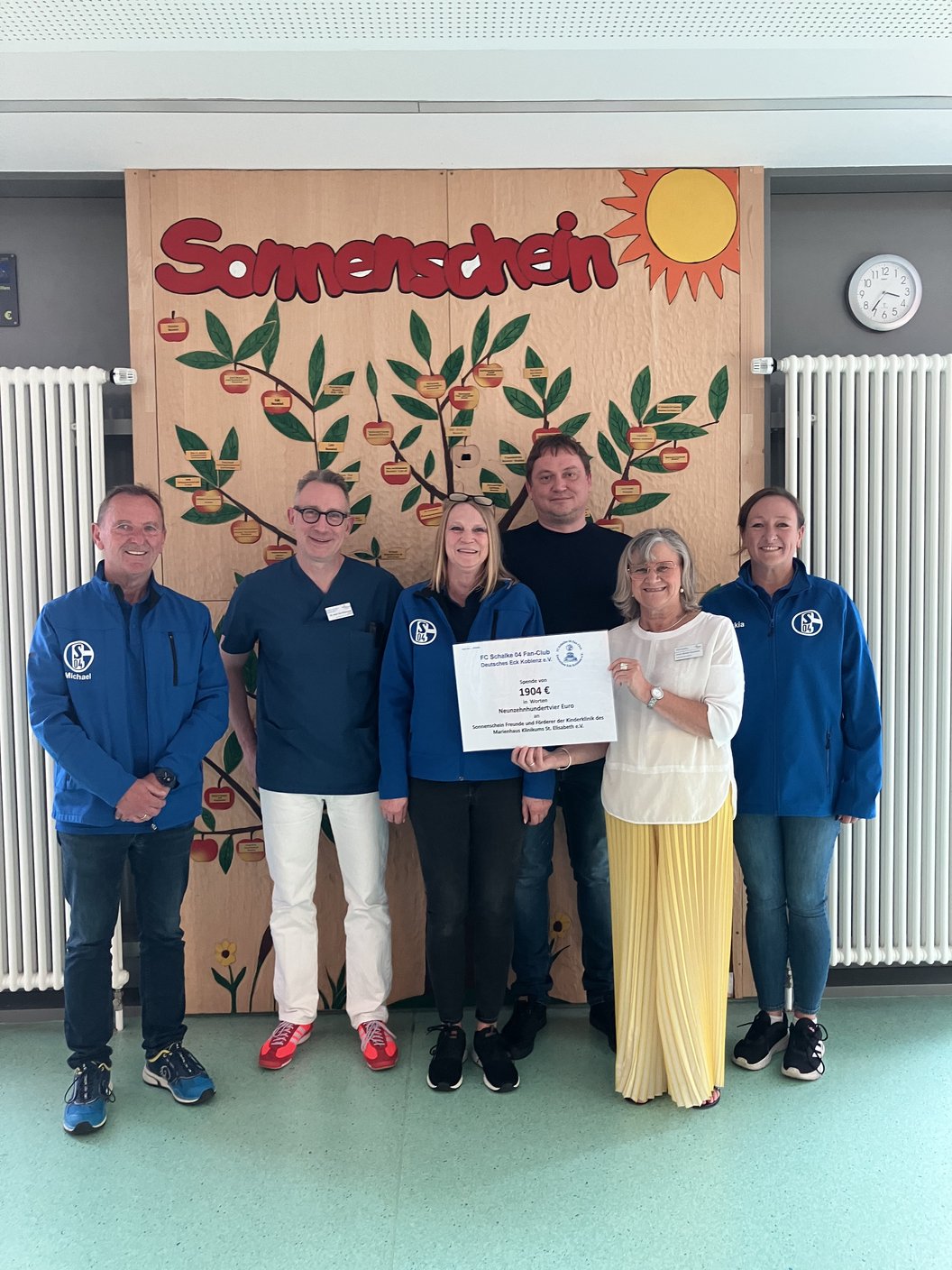 Sechs Personen stehen vor einer Wand mit einem großen Poster, das eine Pflanze und die Aufschrift 'Sonnenschein' zeigt, zwei Personen halten ein großes Schild
