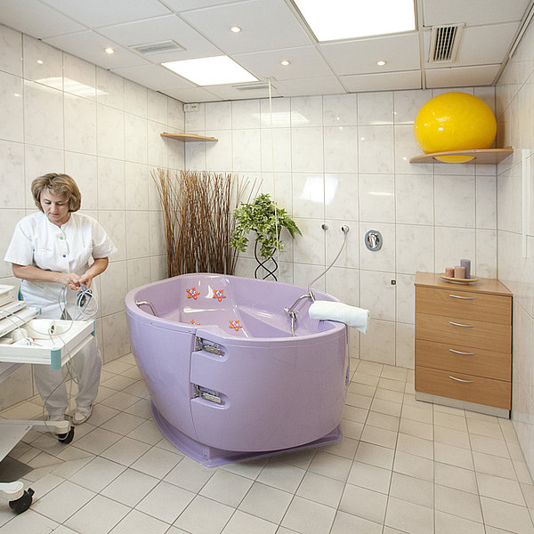 Geburtszimmer mit lila Geburtswanne, medizinischem Gerät auf Rollen und Holzkommode mit gelbem Gymnastikball