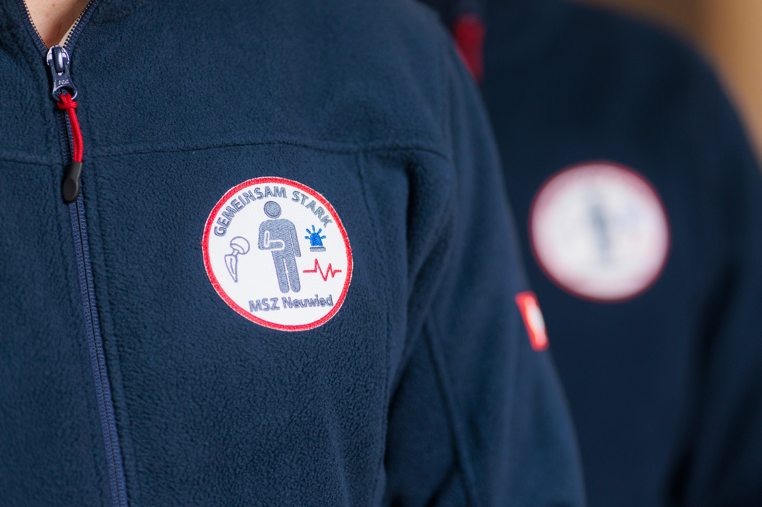 Zwei Personen in dunkelblauen Fleecejacken mit runden Aufnähern, auf denen ein stilisierter Mensch, ein Herzschlag-Symbol und der Text 'GEMEINSAM STARK MSZ Neuwied' zu sehen sind