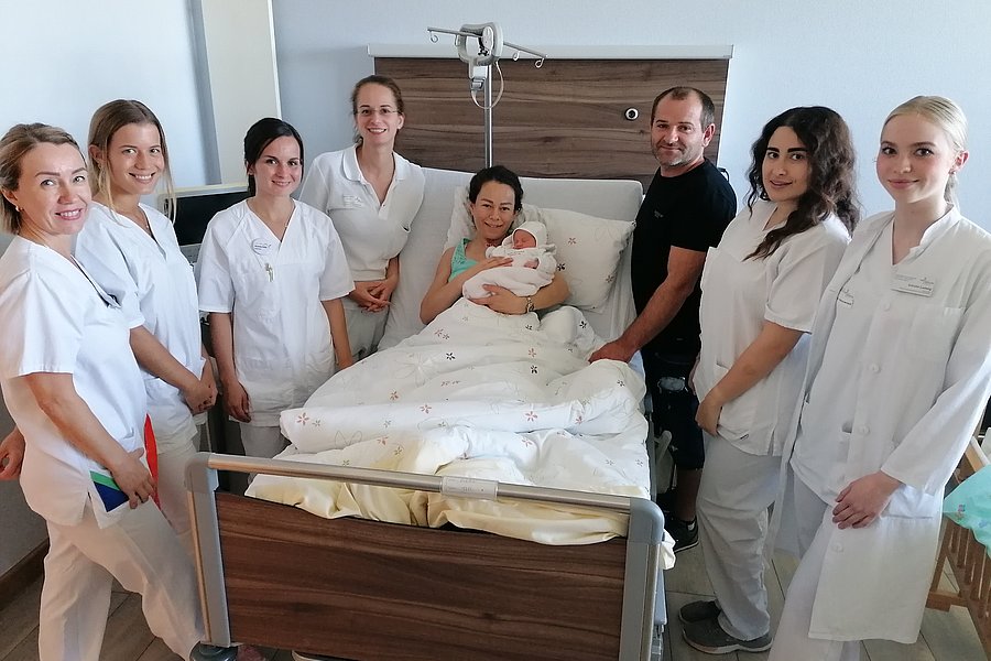 Eine Gruppe von neun Personen in weißen Kitteln steht um ein Bett im Krankenhaus. Eine Person hält ein Baby, während die anderen lächeln und in Richtung der Kamera schauen.