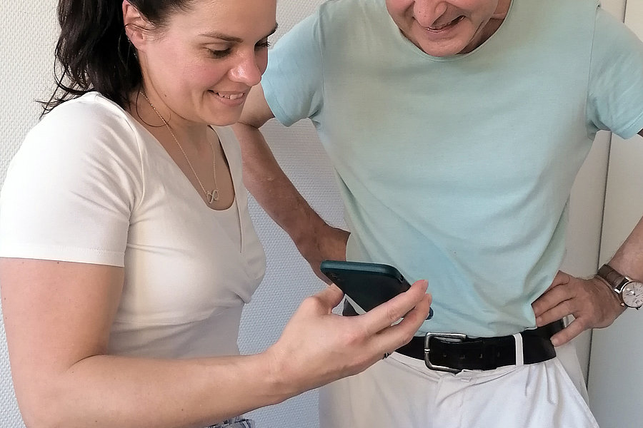Eine Person mit einem weißen Oberteil und einer gestreiften Hose hält ein Smartphone in der Hand. Eine andere Person trägt ein hellblaues T-Shirt und hat die Hände in die Hüften gestützt.