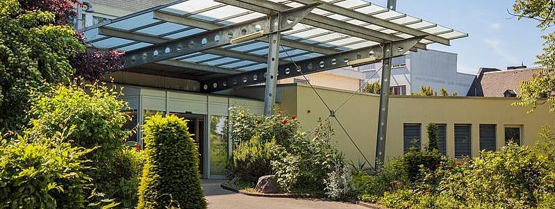 Eingangsbereich eines modernen Gebäudes mit Glasvordach, umgeben von grünen Büschen und Bäumen.
