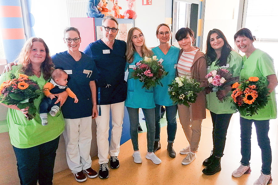 Gruppe von sieben Personen in Arbeitskleidung, die Blumensträuße halten. Eine Person hält ein Baby. Die Kleidung ist grün, blau und weiß.